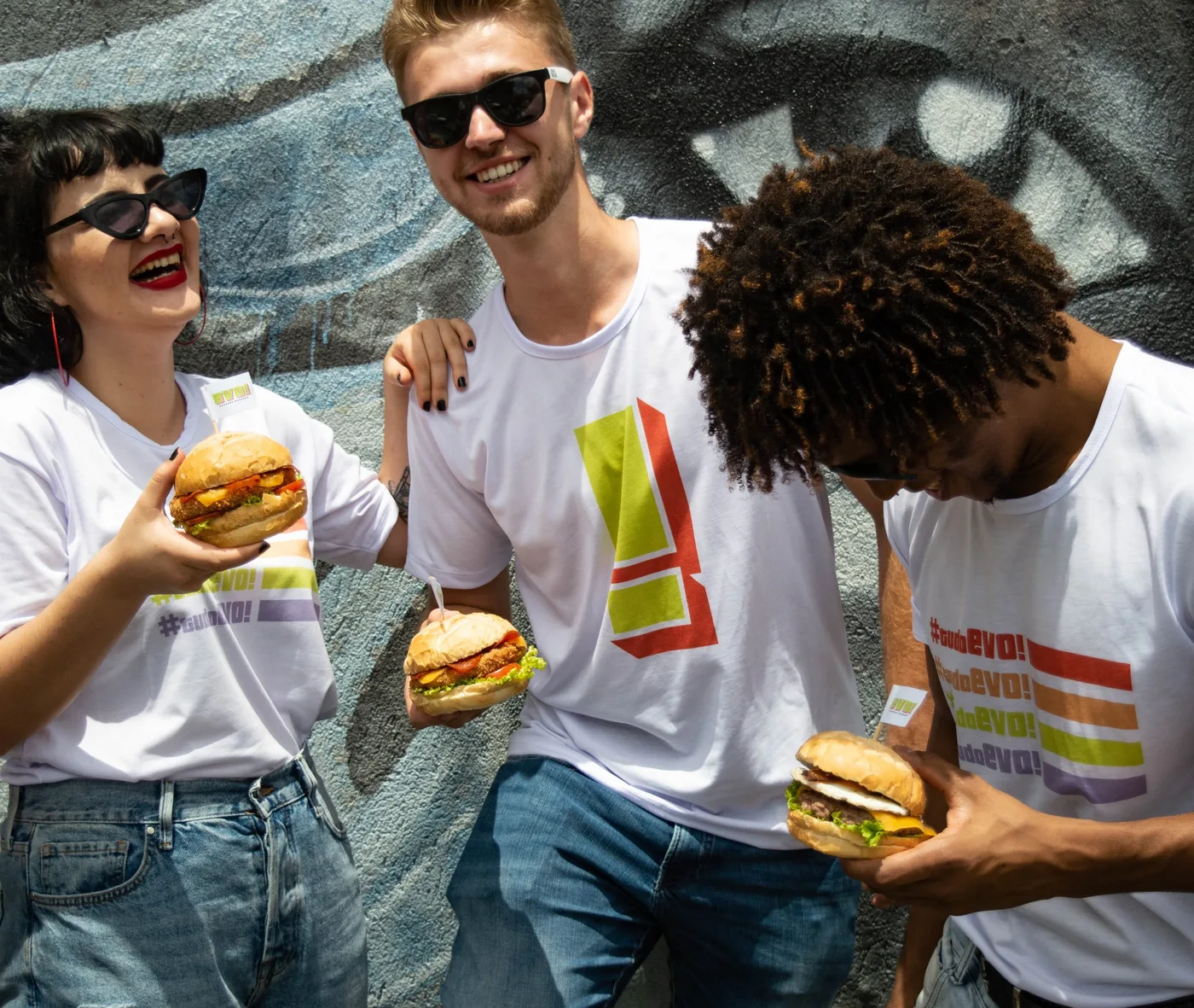 Três amigos sorrindo com camisetas da EVO e hambúrgueres nas mãos, diante de um mural