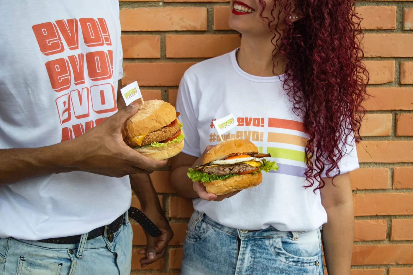 Duas pessoas com camisetas da EVO segurando hambúrgueres com bandeirinhas da marca, diante de uma parede de tijolos