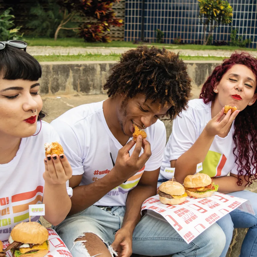 Três amigos com camisetas da EVO comendo bolinhos e hambúrgueres ao ar livre