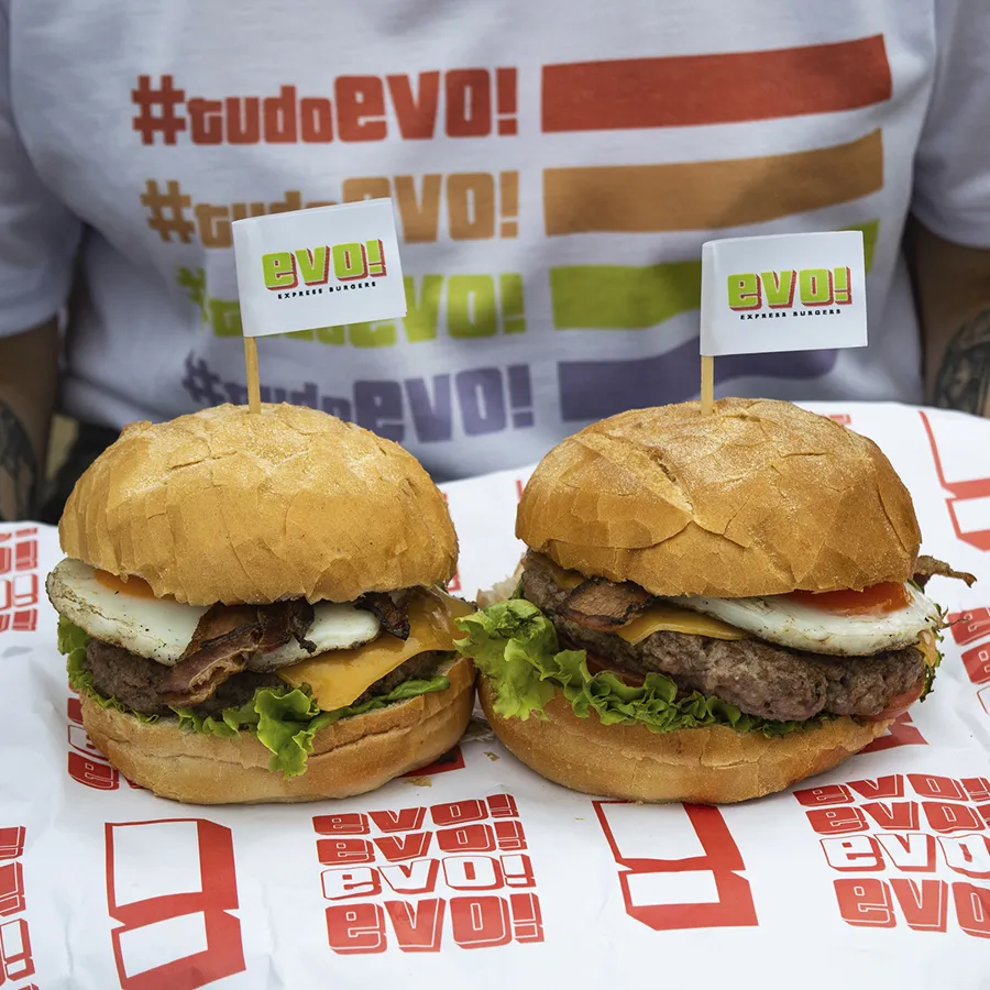 Dois hambúrgueres da EVO sobre papel americano estampado, diante de uma camiseta #tudoEVO