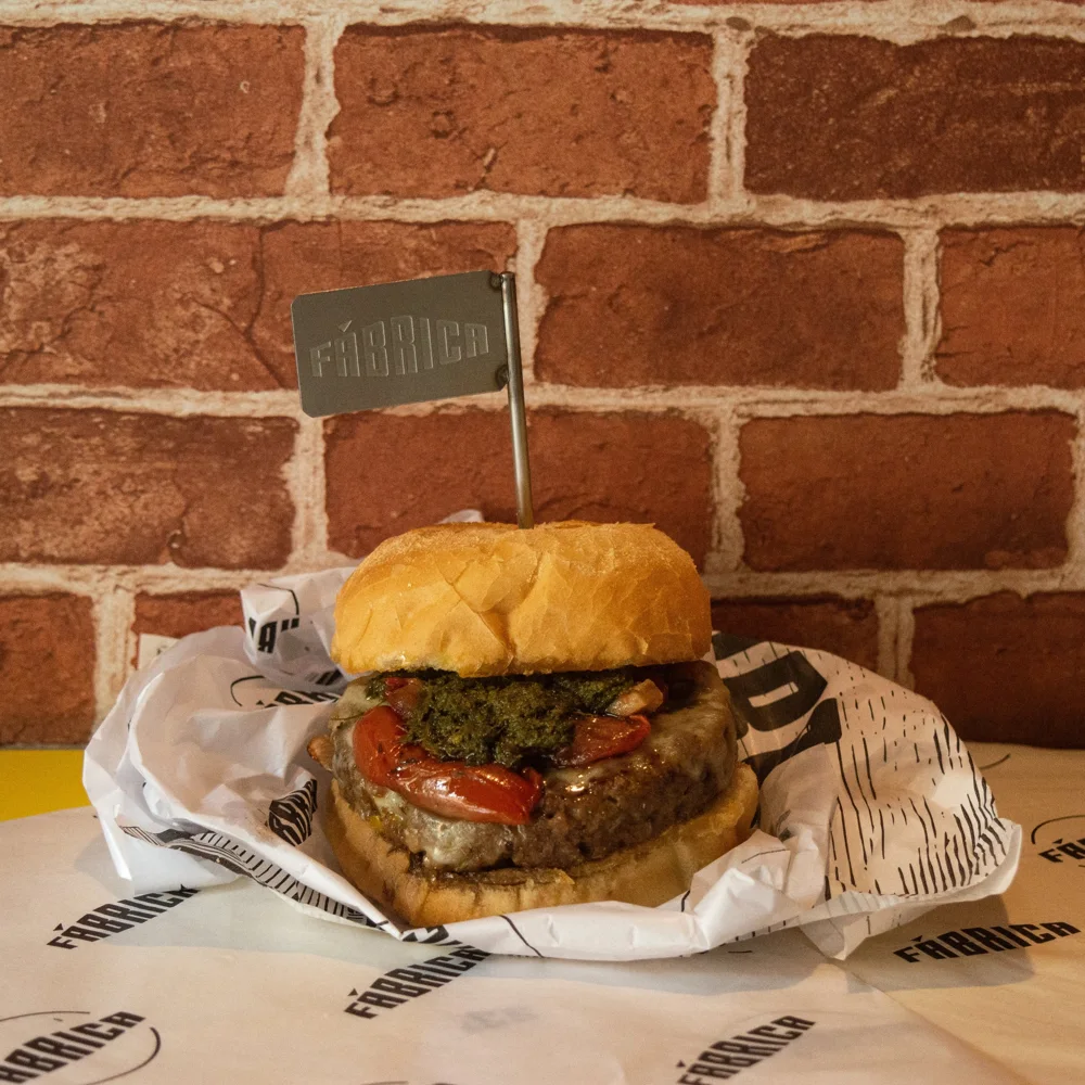 Hambúrguer da Fábrica com bandeirinha de metal, molho verde e tomate assado, sobre papel com a marca, diante de parede de tijolos