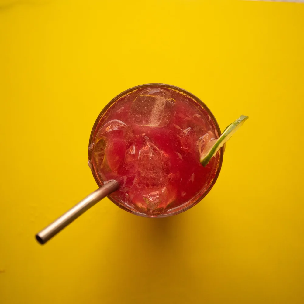 Drink vermelho com gelo, canudo de metal e fatia de limão, visto de cima sobre fundo amarelo