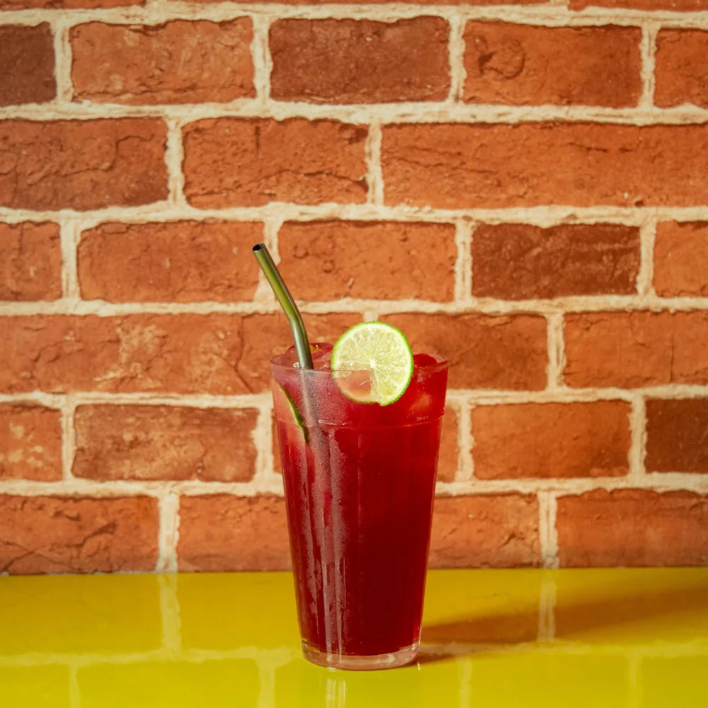 Drink vermelho com canudo de metal e rodela de limão, sobre mesa amarela diante de parede de tijolos