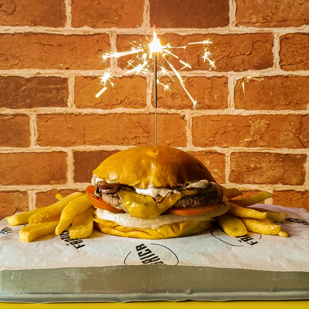 Hambúrguer com batatas fritas e uma vela de faísca acesa, sobre papel da Fábrica, diante de parede de tijolos