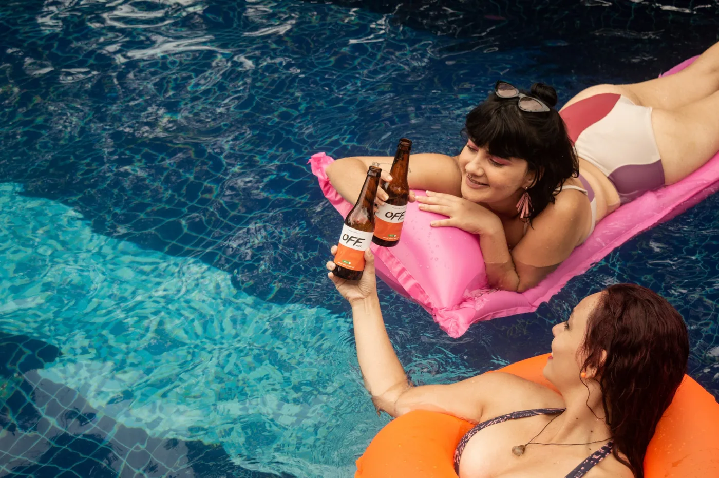 Duas mulheres brindando com garrafas Off Beer numa piscina, vistas de cima, uma deitada em colchão rosa e outra numa boia laranja