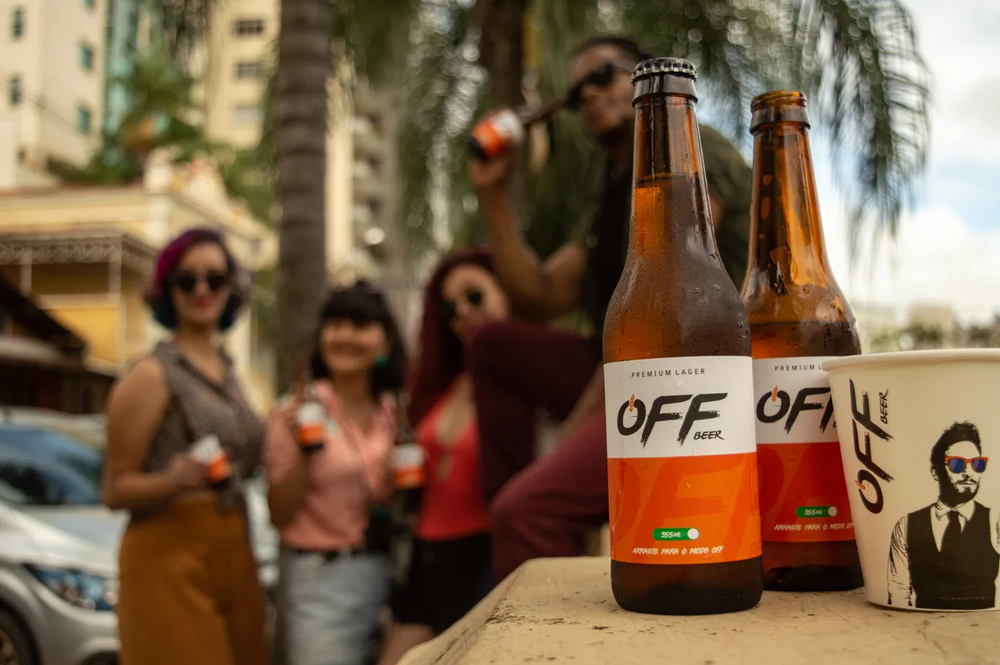 Garrafas e copo Off Beer em primeiro plano sobre um muro, com amigos desfocados ao fundo numa rua com palmeiras