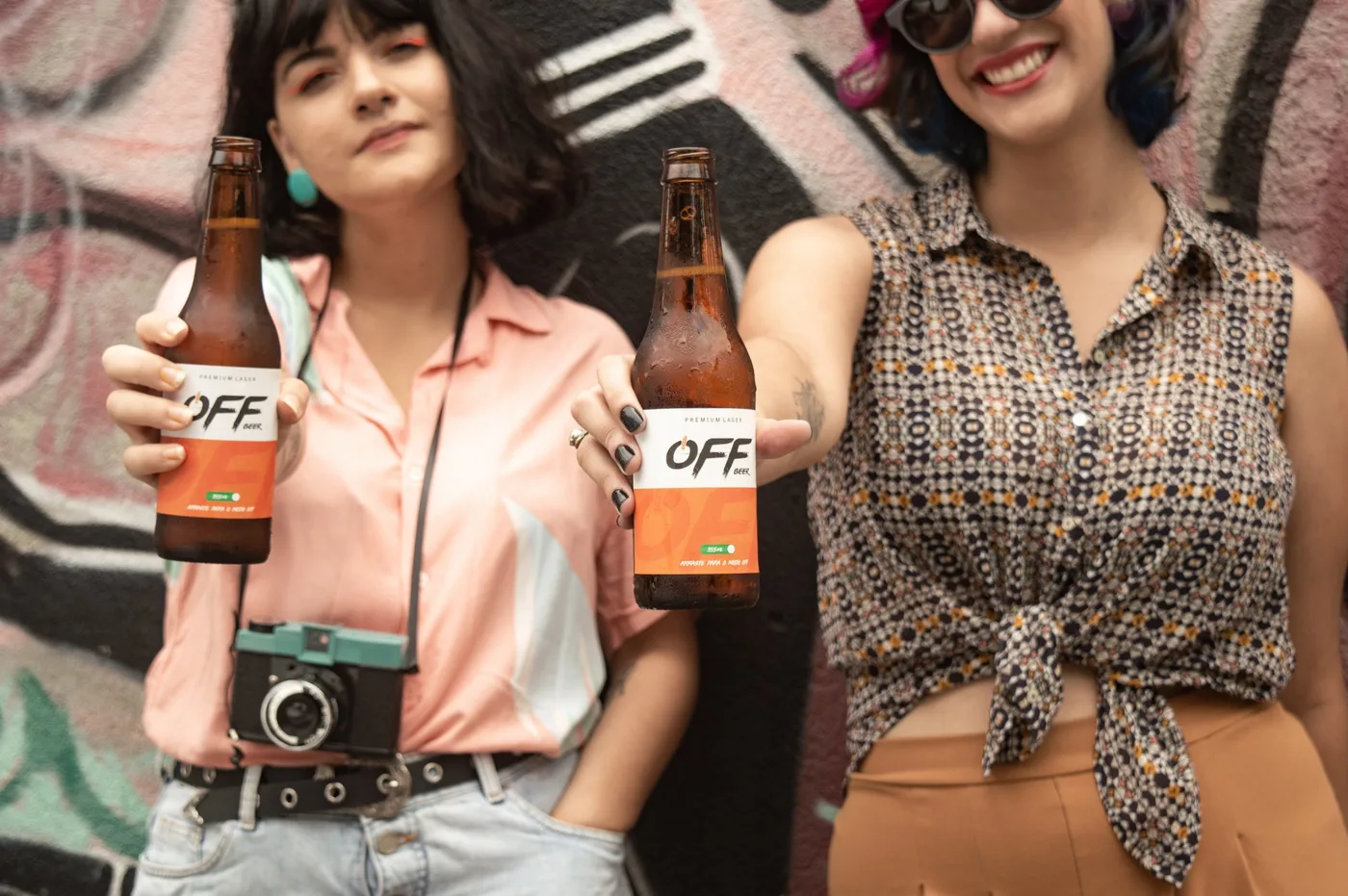Duas mulheres estendendo garrafas Off Beer para a câmera diante de um muro de grafite